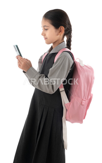 مفهوم التعليم الالكتروني عبر الجوال, متابعة الدروس والواجبات عن بعد, بورتريه من الجانب لطالبة عربية خليجية إماراتية ترتدي المريول المدرسي وحقيبة الظهر تمسك الجوال, تلميذة سعودية تقوم بالبحث عن معلومات لحل الواجبات المدرسية, خلفية بيضاء