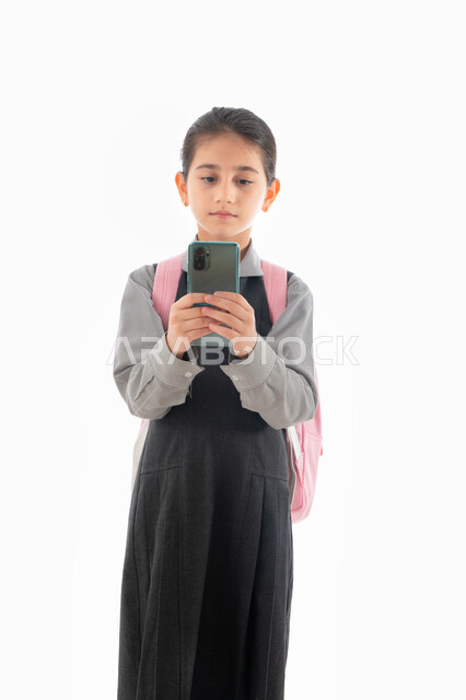 متابعة الدروس والواجبات عن بعد, مفهوم التعليم الالكتروني عبر الجوال, بورتريه لتلميذة عربية خليجية إماراتية ترتدي اليونيفورم المدرسي وحقيبة الظهر تمسك الهاتف المحمول, طالبة سعودية تقوم بالبحث عن معلومات لحل الواجبات المدرسية، خلفية بيضاء