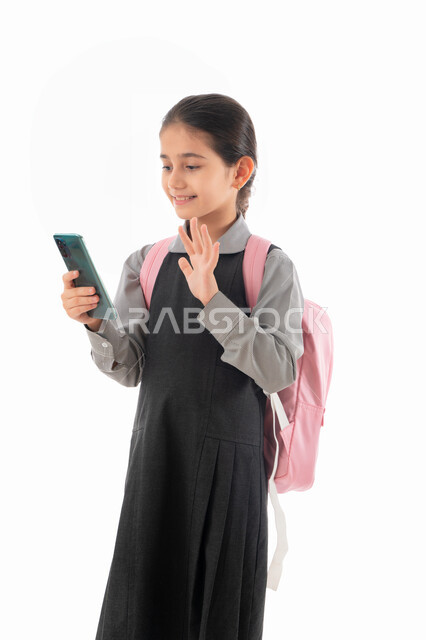 عام دراسي جديد ومخطط تعليمي حديث, بورتريه لطالبة عربية خليجية إمارتية مبتسمة ترتدي المريول المدرسي وحقيبة الظهر وتمسك الهاتف المحمول, تلميذة سعودية تقوم بالتواصل الاجتماعي وإجراء مكالمة فيديو عبر الجوال, استخدام الأجهزة المتطورة لدى الأطفال, خلفية بيضاء