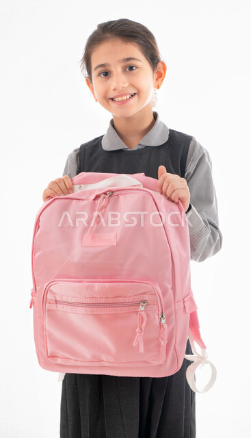 التعليم الحديث في الامارات, الإستعداد للعام الدراسي الجديد، بورتريه لطالبة عربية خليجية اماراتيه ترتدي الزي المدرسي وتحمل بيدها حقيبه الظهر المدرسية، فتاه سعوديه تبتسم وتنظر إلى الكاميرا, إيماءات تدل على الحماس والسعادة لعودة المدارس، خلفية بيضاء.