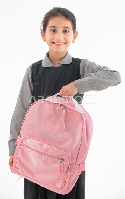Preparing for the new school year, a portrait of an Arab Gulf Emirati student wearing a school uniform and holding a school backpack in her hand, a Saudi girl smiling and looking at the camera, gestures indicating enthusiasm and happiness for the return o