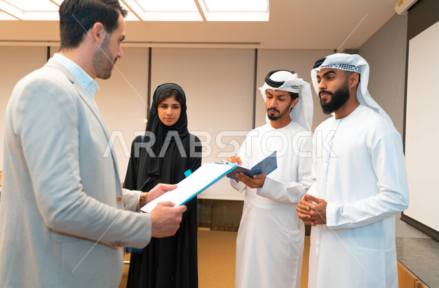 مدرب  يشرح لطلاب وطالبات عرب خليجيين و إماراتين  المحاضره, مدرب جامعي,طلاب إماراتيون يقفنا أمام بروفيسور جامعة ويتم   تدوين الملاحظات على جهاز الحاسب و دفتر الملاحظات , التعليم الجامعي في الإمارات العربية المتحدة , فريق عمل عربي إماراتي  خليجي