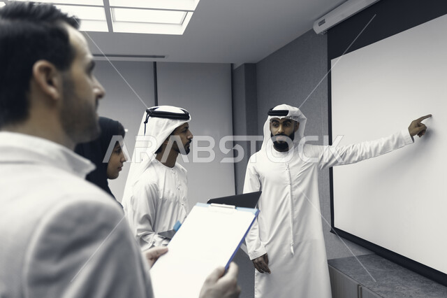 A black and white picture of an Arab Emirati Gulf university professor explaining a lecture to his students, Emirati students standing in front of the professor and taking notes on the computer, an Arab Emirati Gulf work team, meeting at the workplace, di