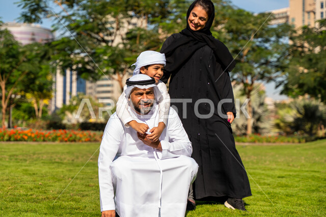 عائله عربيه إماراتية خليجيه مكونه من ثلاث أفراد, أب إماراتي يحمل إبنه خلف ظهره , قضاء وقت ممتع في الحديقه, رحله عائليه, مفهوم الأسره السعيده, إجازه صيفيه, الاستمتاع باللعب الجماعي في المنتزه, تسليه وترفيه