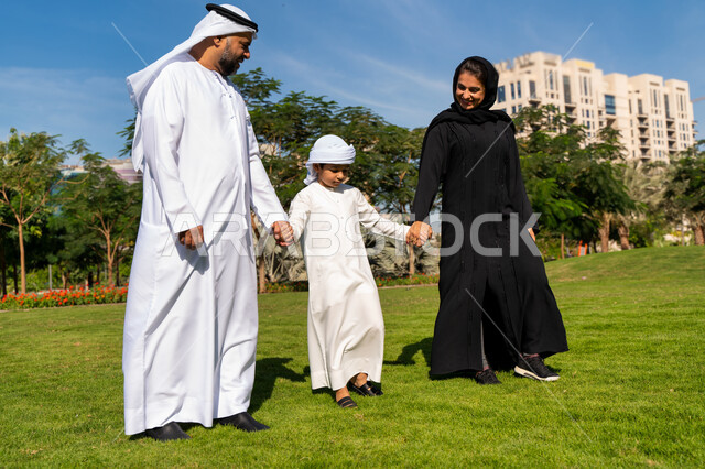 عائله عربيه إماراتية خليجيه مكونه من ثلاث أفراد, أب وأم إماراتيان يمسكان يد طفلهم, قضاء وقت ممتع في الحديقه , رحله عائليه, مفهوم الأسره السعيده, إجازه صيفيه, الاستمتاع باللعب الجماعي في المنتزه, تسليه وترفيه