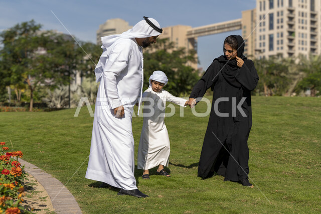 عائله عربيه إماراتية خليجيه مكونه من ثلاث أفراد, أب وأم إماراتيان يمسكان يد طفلهم, قضاء وقت ممتع في الحديقه , رحله عائليه, مفهوم الأسره السعيده, إجازه صيفيه, الاستمتاع باللعب الجماعي في المنتزه, تسليه وترفيه