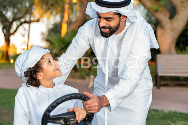 أب عربي خليجي إماراتي يرتدي الزي التقليدي يلعب مع ابنه في الحديقه ، اللعب بالدباب الكهربائي, مفهوم الأسرة السعيدة ، قضاء أوقات عائلية ممتعة, رحله عائليه في الصيف, اجازه صيفيه سعيده, عطلة نهاية الاسبوع, اللعب مع الابناء