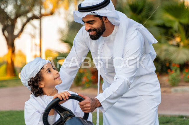أب عربي خليجي إماراتي يرتدي الزي التقليدي يلعب مع ابنه في الحديقه ، اللعب بالدباب الكهربائي, مفهوم الأسرة السعيدة ، قضاء أوقات عائلية ممتعة, رحله عائليه في الصيف, اجازه صيفيه سعيده, عطلة نهاية الاسبوع, اللعب مع الابناء