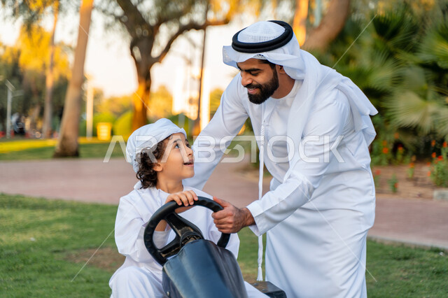أب عربي خليجي إماراتي يرتدي الزي التقليدي يلعب مع ابنه في الحديقه ، اللعب بالدباب الكهربائي, مفهوم الأسرة السعيدة ، قضاء أوقات عائلية ممتعة, رحله عائليه في الصيف, اجازه صيفيه سعيده, عطلة نهاية الاسبوع, اللعب مع الابناء