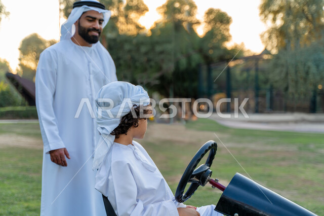 أب عربي خليجي إماراتي يرتدي الزي التقليدي يلعب مع ابنه في الحديقه ، اللعب بالدباب الكهربائي, مفهوم الأسرة السعيدة ، قضاء أوقات عائلية ممتعة, رحله عائليه في الصيف, اجازه صيفيه سعيده, عطلة نهاية الاسبوع, اللعب مع الابناء