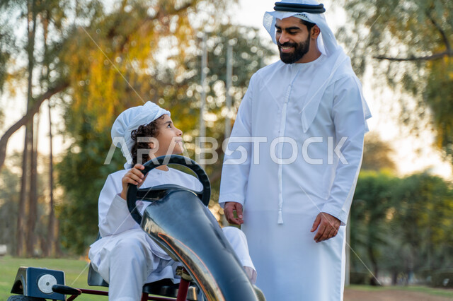أب عربي خليجي إماراتي يرتدي الزي التقليدي يلعب مع ابنه في الحديقه ، اللعب بالدباب الكهربائي, مفهوم الأسرة السعيدة ، قضاء أوقات عائلية ممتعة, رحله عائليه في الصيف, اجازه صيفيه سعيده, عطلة نهاية الاسبوع, اللعب مع الابناء