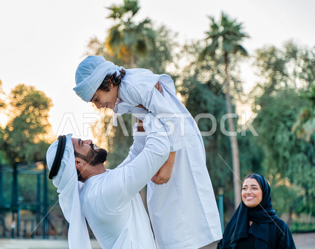 عائله عربيه إماراتيه  خليجيه مكونه من ثلاث أفراد, أب إماراتي يرتدي الزي التقليدي يلعب مع ابنه و يرفعه عاليا , رحله عائليه عند غروب الشمس , مفهوم الأسره السعيده, إجازه صيفيه, الاستمتاع باللعب الجماعي في المنتزه, تسليه وترفيه, عطلة نهاية الاسبوع