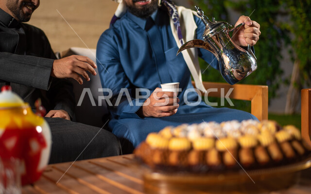 A close-up of a group of Saudi Gulf Arab youth spending fun times in one of the tourist chalets in Saudi Arabia, a young man pouring coffee, a winter atmosphere, a happy youth atmosphere, a session with people and acquaintance, a meeting of friends, a you