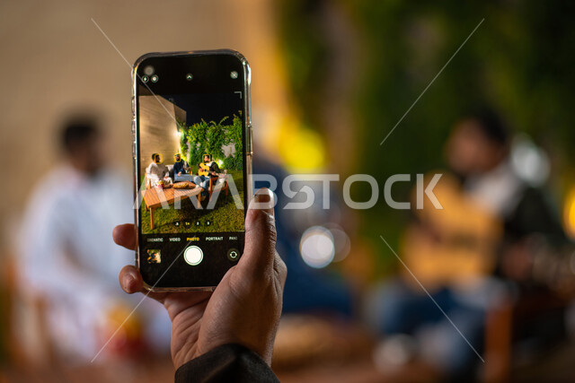 صورة مقربة لشاب يمسك بالهاتف المحمول ويقوم بتصوير اصدقائه، مجموعة من الشباب العرب الخليجيين السعوديين يقضون أوقات ممتعة في إحدى الشاليهات السياحية بالسعودية، التقاط الصور التذكارية، أجواء شتوية، جلسة وناسة وتعارف، إجتماع الأصدقاء، سهرة شبابية