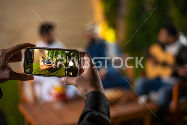 صورة مقربة لشاب يمسك بالهاتف المحمول ويقوم بتصوير اصدقائه، مجموعة من الشباب العرب الخليجيين السعوديين يقضون أوقات ممتعة في إحدى الشاليهات السياحية بالسعودية، التقاط الصور التذكارية، أجواء شتوية، جلسة وناسة وتعارف، إجتماع الأصدقاء، سهرة شبابية