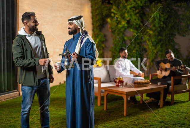 A close-up of a group of Saudi Gulf Arab youth spending fun times in one of the tourist chalets in Saudi Arabia, a young man pouring coffee, a winter atmosphere, a happy youth atmosphere, a session with people and acquaintance, a meeting of friends, a you