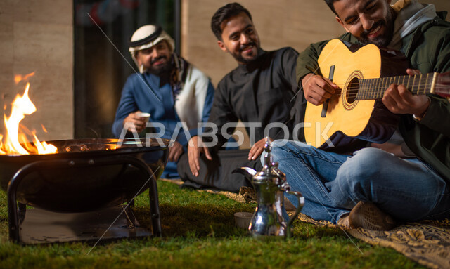 A close-up of a group of Saudi Gulf Arab youth spending fun times in one of the tourist chalets in Saudi Arabia, youth entertainment activities, a young man playing the harp, winter atmosphere, a happy youth atmosphere, a session, people and acquaintance,