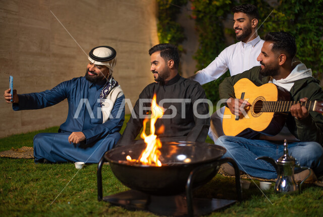 مجموعة من الشباب العرب الخليجيين السعوديين يقضون أوقات ممتعة في إحدى الشاليهات السياحية بالسعودية، شاب يعزف على آلة القيثارة، أجواء شتوية،  التقاط صور السيلفي التذكارية من خلال الهاتف المحمول، جلسة وناسة وتعارف، إجتماع الأصدقاء، سهرة شبابية