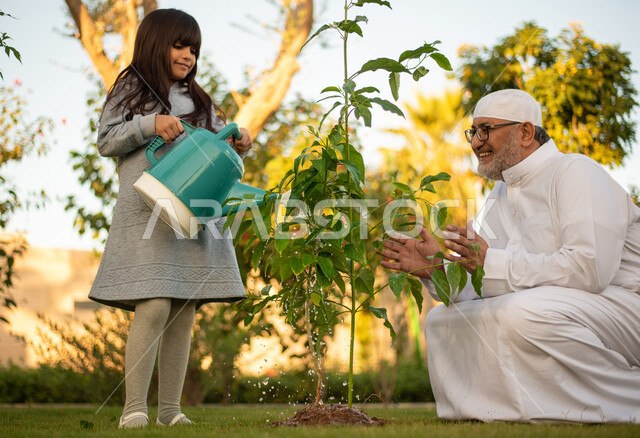 رجل عربي خليجي سعودي كبير بالسن يقضي وقت ممتع مع حفيدته في ري الأشجار بإستخدام وعاء السقي ، فعاليات وأجواء ترفيهية ، تعابير تدل على السعادة ، خلفية الطبيعة ، الأِشجار الطبيعية الخضراء ، التسليه والترفيه ، مشاركة عائلية