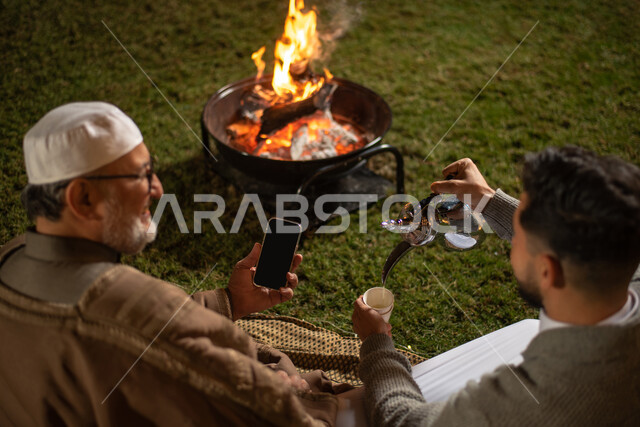 زاوية من الخلف لرجل عربي خليجي سعودي مع رجل كبير بالسن في إحدى الشاليهات السياحية بالسعودية، إستخدام الهاتف المحمول، صب القهوة، الحوارات والنقاشات، أجواء عائلية سعيدة، أجواء شتوية، منتجعات وشاليهات ترفيهية بالسعودية، سهرة رجالية