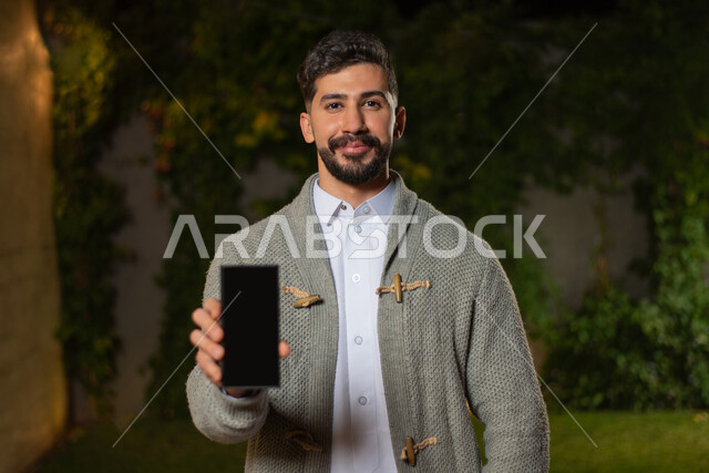 صورة مقربة لرجل عربي خليجي سعودي ، مبتسم ينظر إلى الكاميرا ، عرض شاشة سوداء بإستخدام الهاتف المحمول ، تعابير وجه تدل على السعادة ، أزياء الشتاء ، ملابس شتوية ، خلفية الطبيعة