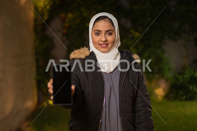 صورة مقربة لمرأة عربية خليجية سعودية ، مبتسمة تنظر إلى الكاميرا ، عرض شاشة سوداء بإستخدام الهاتف المحمول ، تعابير وجه تدل على السعادة ، أزياء الشتاء ، ملابس شتوية ، خلفية الطبيعة