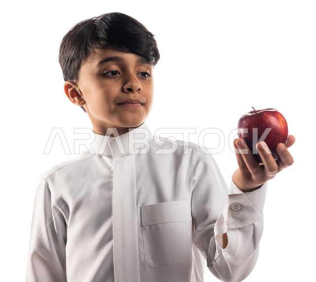 Portrait of a Saudi Arabian Gulf boy looking at the apple fruit, the concept of health, maintaining a healthy body, a healthy morning breakfast, a healthy diet, fresh and healthy apple fruit, white background