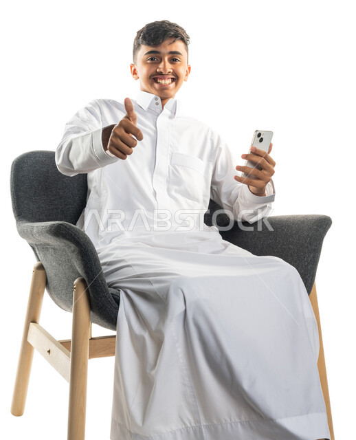 بورتريه لولد عربي سعودي خليجي مبتسم ، يجلس على كرسي ، إستخدام الهاتف المحمول ، يشير بإصبعه الإبهام لأعلى ، تعابير تدل على الإعجاب ، التواصل مع الإصدقاء ، تصفح الإنترنت ، إستخدام وسائل التواصل الإجتماعي ، خلفية بيضاء
