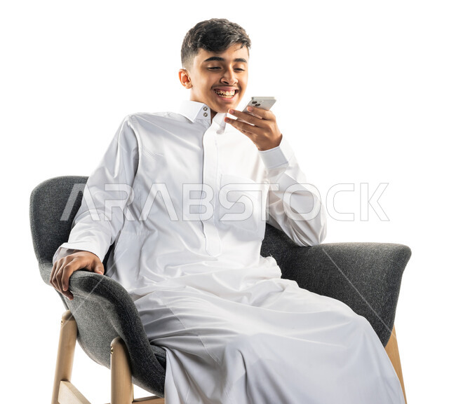 بورتريه لولد عربي سعودي خليجي مبتسم ، يجلس على كرسي ، إستخدام الهاتف المحمول ، التواصل مع الإصدقاء ، تصفح الإنترنت ، إستخدام وسائل التواصل الإجتماعي ، خلفية بيضاء