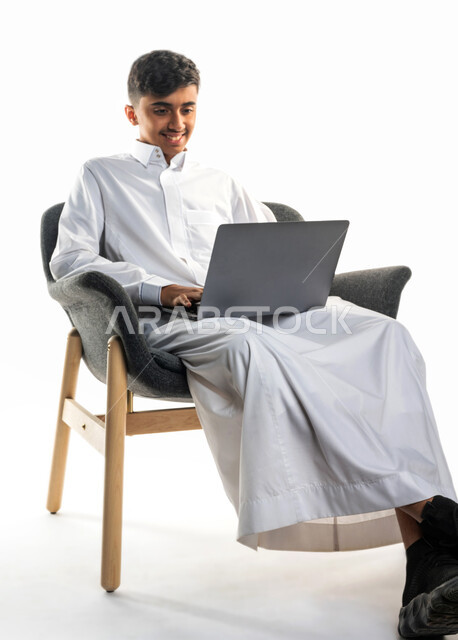 بورتريه لولد عربي سعودي خليجي ، يجلس على كرسي ،   إستخدام جهاز الحاسوب المحمول ، التعليم الإلكتروني ، التعليم عن بعد ، تصفح الإنترنت ، محادثات ودردشة ، خلفية بيضاء