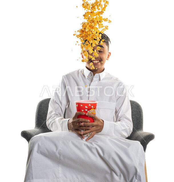بورتريه لولد عربي سعودي خليجي، يجلس على كرسي، يحمل بيده دلو الفشار، حبات فشار متطايرة، مشاهدة الأفلام والعروض المسرحية، تعابير تدل على السعادة، أنشطة ترفيهية، أفلام وسينما، قضاء أوقات ممتعة، خلفية بيضاء