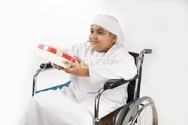 بورتريه لطفل عربي إماراتي خليجي يرتدي الزي التقليدي من ذوي الهمم يجلس على كرسي متحرك و يمسك بيده علبة هدية   , التمكين الاجتماعي, اهتمام ودعم المجتمع الإماراتي لذوي الاحتياجات الخاصه, تقديم الحب و الرعاية و الاهتمام , خلفية بيضاء 