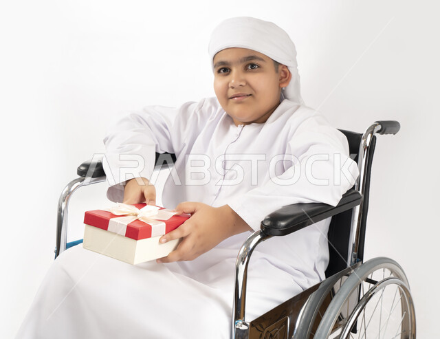 بورتريه لطفل عربي إماراتي خليجي يرتدي الزي التقليدي من ذوي الهمم يجلس على كرسي متحرك و يمسك بيده علبة هدية   , التمكين الاجتماعي, اهتمام ودعم المجتمع الإماراتي لذوي الاحتياجات الخاصه, تقديم الحب و الرعاية و الاهتمام , خلفية بيضاء 