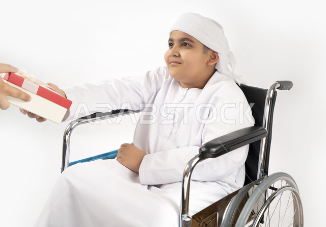 بورتريه لطفل عربي إماراتي خليجي يرتدي الزي التقليدي من ذوي الهمم يجلس على كرسي متحرك و يمسك بيده علبة هدية   , التمكين الاجتماعي, اهتمام ودعم المجتمع الإماراتي لذوي الاحتياجات الخاصه, تقديم الحب و الرعاية و الاهتمام , خلفية بيضاء 