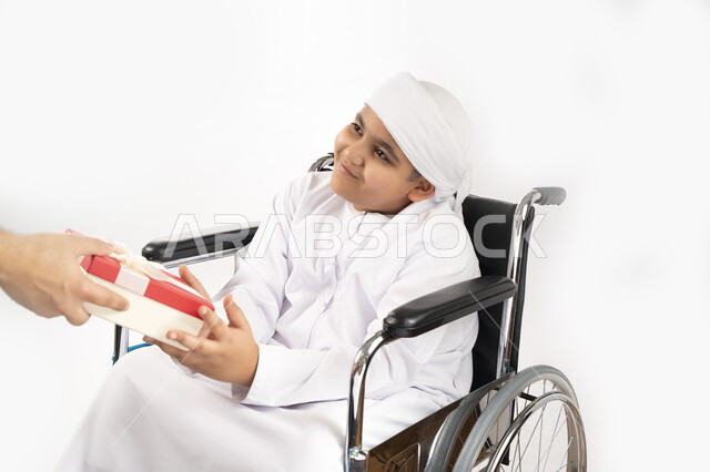 بورتريه لطفل عربي إماراتي خليجي يرتدي الزي التقليدي من ذوي الهمم يجلس على كرسي متحرك و يمسك بيده علبة هدية   , التمكين الاجتماعي, اهتمام ودعم المجتمع الإماراتي لذوي الاحتياجات الخاصه, تقديم الحب و الرعاية و الاهتمام , خلفية بيضاء 