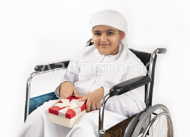 بورتريه لطفل عربي إماراتي خليجي يرتدي الزي التقليدي من ذوي الهمم يجلس على كرسي متحرك و يمسك بيده علبة هدية   , التمكين الاجتماعي, اهتمام ودعم المجتمع الإماراتي لذوي الاحتياجات الخاصه, تقديم الحب و الرعاية و الاهتمام , خلفية بيضاء 