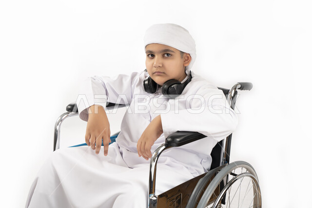 صورة مقربة لبورتريه لطفل عربي إماراتي خليجي يرتدي الزي التقليدي من ذوي الهمم يجلس على كرسي متحرك  يرتدي الزي التقليدي ويضع السماعة على رقبته , التمكين الاجتماعي, اهتمام ودعم المجتمع الإماراتي لذوي الاحتياجات الخاصه, تقديم الحب و الرعاية و الاهتمام , خلفية