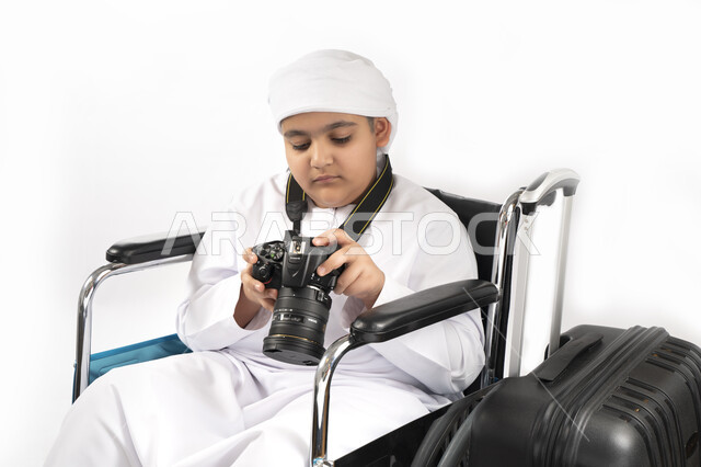 صورة مقربة لبورتريه طفل عربي إماراتي خليجي يرتدي الزي التقليدي من ذوي الهمم يجلس على كرسي متحرك يمسك بيده كاميرا عالية الجودة و بجانبه شنطة السفر   , التمكين الاجتماعي, اهتمام ودعم المجتمع الإماراتي لذوي الاحتياجات الخاصه, تقديم الحب و الرعاية و الاهتمام,