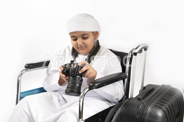 صورة مقربة لبورتريه طفل عربي إماراتي خليجي يرتدي الزي التقليدي من ذوي الهمم يجلس على كرسي متحرك يمسك بيده كاميرا عالية الجودة و بجانبه شنطة السفر   , التمكين الاجتماعي, اهتمام ودعم المجتمع الإماراتي لذوي الاحتياجات الخاصه, تقديم الحب و الرعاية و الاهتمام,