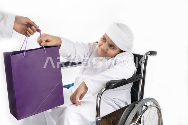 بورتريه لطفل عربي إماراتي خليجي يرتدي الزي التقليدي من ذوي الهمم يجلس على كرسي متحرك و يمسك بيده علبة هدية   , التمكين الاجتماعي, اهتمام ودعم المجتمع الإماراتي لذوي الاحتياجات الخاصه, تقديم الحب و الرعاية و الاهتمام , خلفية بيضاء 