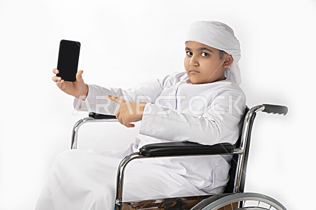 بورتريه لطفل عربي إماراتي خليجي يرتدي الزي التقليدي من ذوي الهمم يجلس على كرسي متحرك و يمسك بيده هاتف محمول بشاشة سوداء مفرغة  , التمكين الاجتماعي, اهتمام ودعم المجتمع الإماراتي لذوي الاحتياجات الخاصه, تقديم الحب و الرعاية و الاهتمام , خلفية بيضاء 