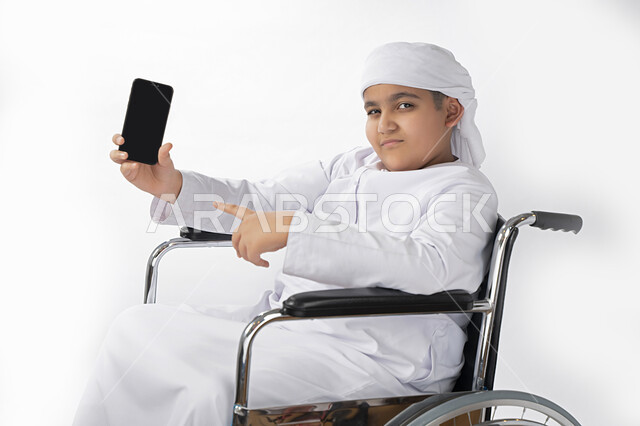 بورتريه لطفل عربي إماراتي خليجي يرتدي الزي التقليدي من ذوي الهمم يجلس على كرسي متحرك و يمسك بيده هاتف محمول بشاشة سوداء مفرغة  , التمكين الاجتماعي, اهتمام ودعم المجتمع الإماراتي لذوي الاحتياجات الخاصه, تقديم الحب و الرعاية و الاهتمام , خلفية بيضاء 