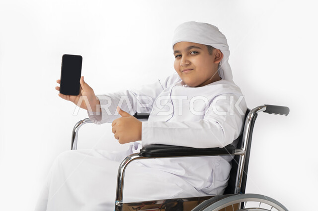 بورتريه لطفل عربي إماراتي خليجي يرتدي الزي التقليدي من ذوي الهمم يجلس على كرسي متحرك و يمسك بيده هاتف محمول بشاشة سوداء مفرغة  , التمكين الاجتماعي, اهتمام ودعم المجتمع الإماراتي لذوي الاحتياجات الخاصه, تقديم الحب و الرعاية و الاهتمام , خلفية بيضاء 