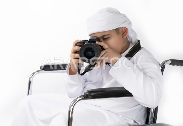 بورتريه لطفل عربي إماراتي خليجي يرتدي الزي التقليدي من ذوي الهمم يجلس على كرسي متحرك ويرتدي الزي التقليدي ويمسك بيده كاميرا تصوير عالية الجودة  , التمكين الاجتماعي, اهتمام ودعم المجتمع الإماراتي لذوي الاحتياجات الخاصه, خلفية بيضاء 