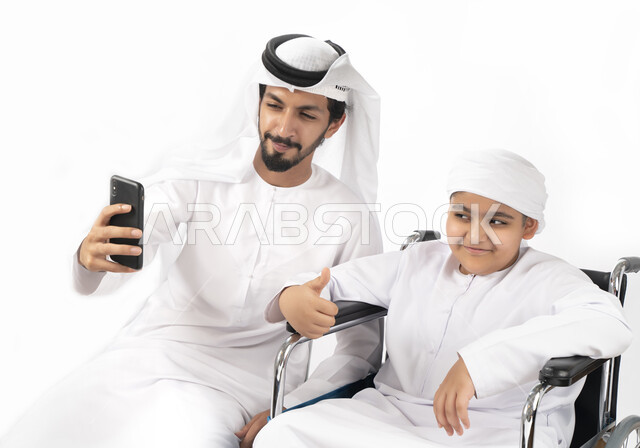 بورتريه لطفل عربي إماراتي خليجي من ذوي الهمم يجلس على كرسي متحرك ويرتدي الزي التقليدي كندورة وعمامه يلتقط صورة سيلفي مع والده  ، التمكين الاجتماعي  ، اهتمام ودعم المجتمع الإماراتي لذوي  الاحتياجات الخاصه، شعور ذوي الهمم بالراحه والطمأنينه في بيئتهم , خلفي