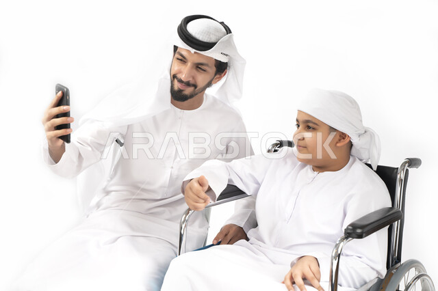 بورتريه لطفل عربي إماراتي خليجي من ذوي الهمم يجلس على كرسي متحرك ويرتدي الزي التقليدي كندورة وعمامه يلتقط صورة سيلفي مع والده  ، التمكين الاجتماعي  ، اهتمام ودعم المجتمع الإماراتي لذوي  الاحتياجات الخاصه، شعور ذوي الهمم بالراحه والطمأنينه في بيئتهم , خلفي