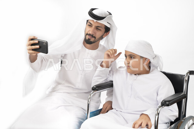بورتريه لطفل عربي إماراتي خليجي من ذوي الهمم يجلس على كرسي متحرك ويرتدي الزي التقليدي كندورة وعمامه يلتقط صورة سيلفي مع والده  ، التمكين الاجتماعي  ، اهتمام ودعم المجتمع الإماراتي لذوي  الاحتياجات الخاصه، شعور ذوي الهمم بالراحه والطمأنينه في بيئتهم , خلفي