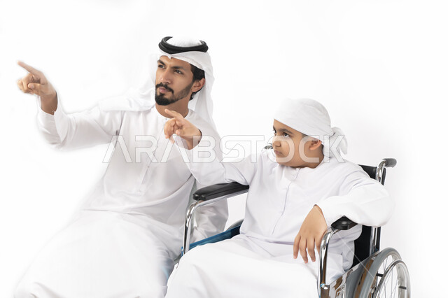 بورتريه لطفل عربي إماراتي خليجي من ذوي الهمم يجلس على كرسي متحرك ويرتدي الزي التقليدي كندورة وعمامه مع والده ويأشران بيدهم الى شيء ما  ، التمكين الاجتماعي  ، اهتمام ودعم المجتمع الإماراتي لذوي  الاحتياجات الخاصه، شعور ذوي الهمم بالراحه والطمأنينه في بيئته