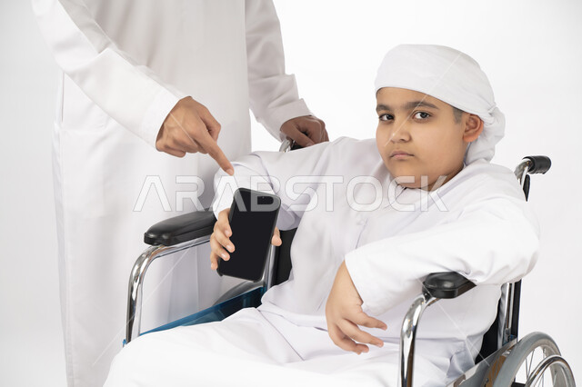 بورتريه لطفل عربي إماراتي خليجي يرتدي الزي التقليدي من ذوي الهمم يجلس على كرسي متحرك و يمسك بيده هاتف محمول بشاشة سوداء مفرغة  , التمكين الاجتماعي, اهتمام ودعم المجتمع الإماراتي لذوي الاحتياجات الخاصه, تقديم الحب و الرعاية و الاهتمام , خلفية بيضاء 