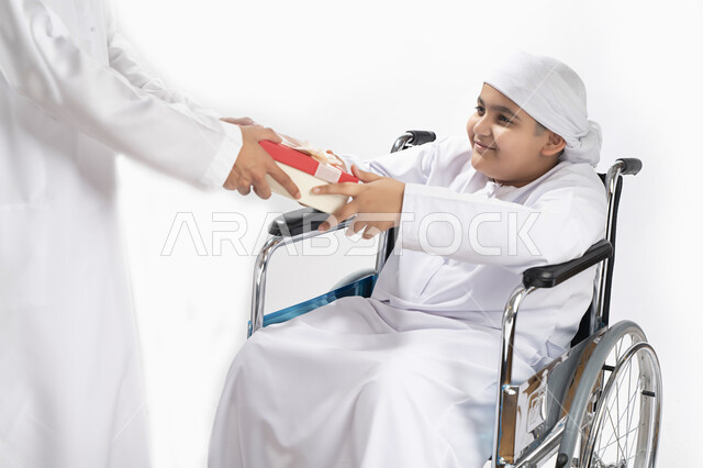 Portrait of an Arab Emirati Gulf child dressed in traditional clothes of people of determination, sitting in a wheelchair and holding a gift box in his hand, social empowerment, the interest and support of the Emirati community for people with special nee